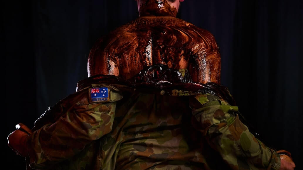 A person with their back turned, covered in a dark, glossy substance, wears a camouflaged jacket featuring an Australian flag patch.