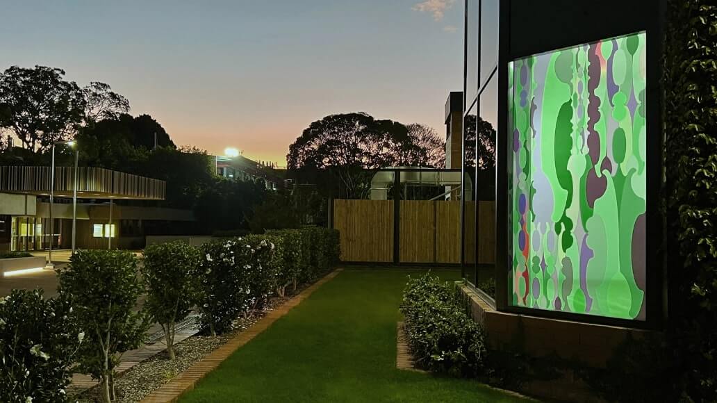 A modern building with a large colorful abstract window design at dusk, surrounded by greenery.