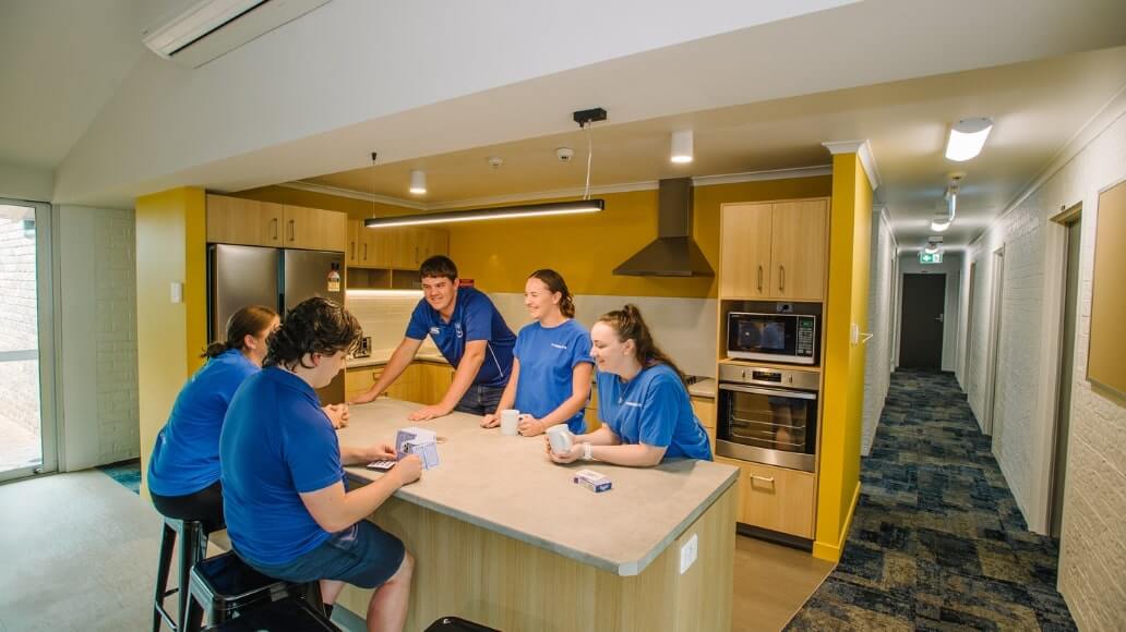 Five people in blue shirts sit and stand around a  student kitchen at Steele Rudd, one of UniSQ's Residential Colleges.