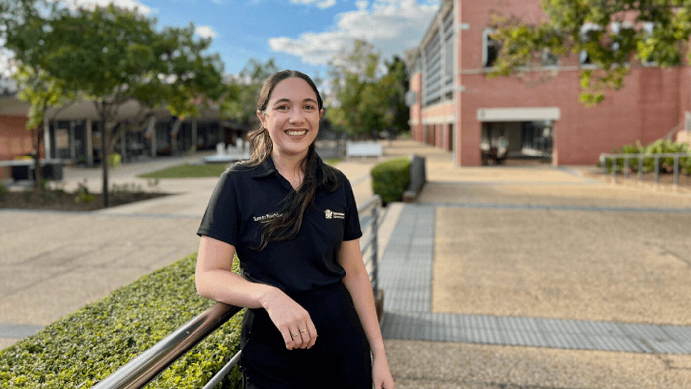 A woman smiles while standing outdoors on a campus walkway, near a hedge and a brick building under a partly cloudy sky.