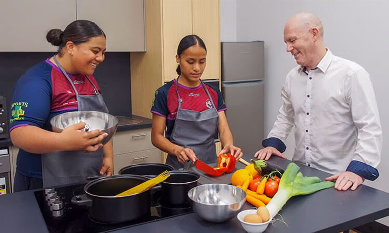 Three people in a kitchen: two wearing aprons, one cutting a bell pepper, with ingredients and cookware on the counter.