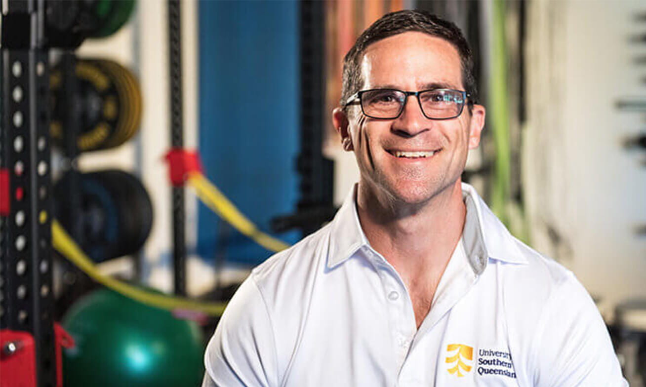 A man wearing glasses and a University of Southern Queensland shirt smiles in a gym setting with exercise equipment in the background.