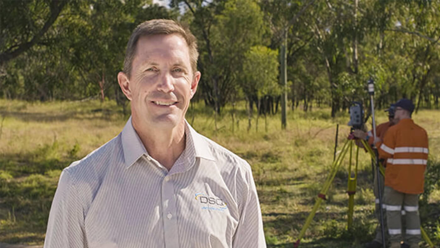 A man in a shirt stands outside smiling, with trees in the background. Another person in an orange jacket is surveying the area using equipment.