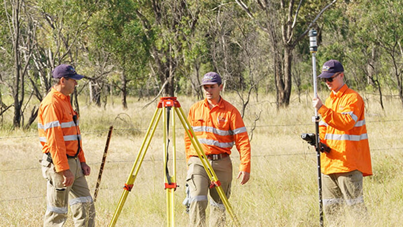 Three people in orange safety uniforms conduct a land survey in a grassy area, using tripods and measuring equipment. Trees are visible in the background.