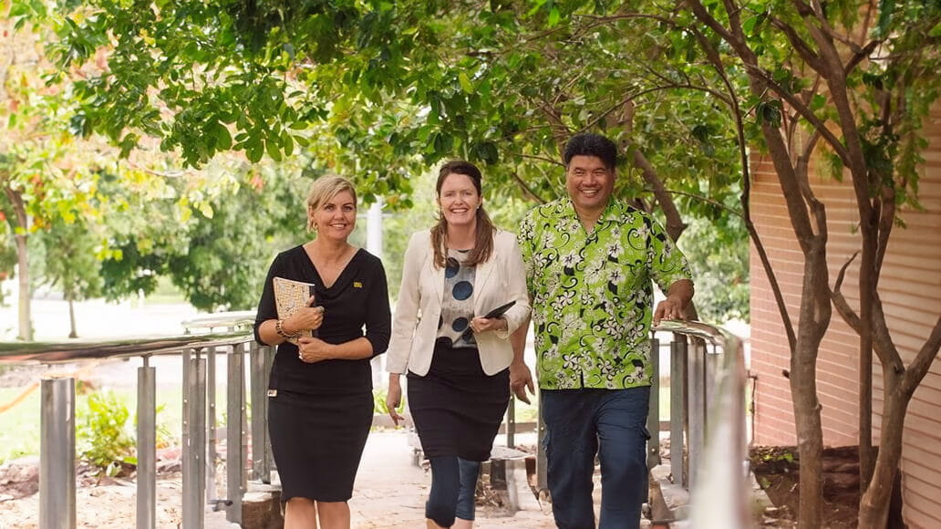 Three people walking on a park walkway, engaged in a conversation, surrounded by greenery.