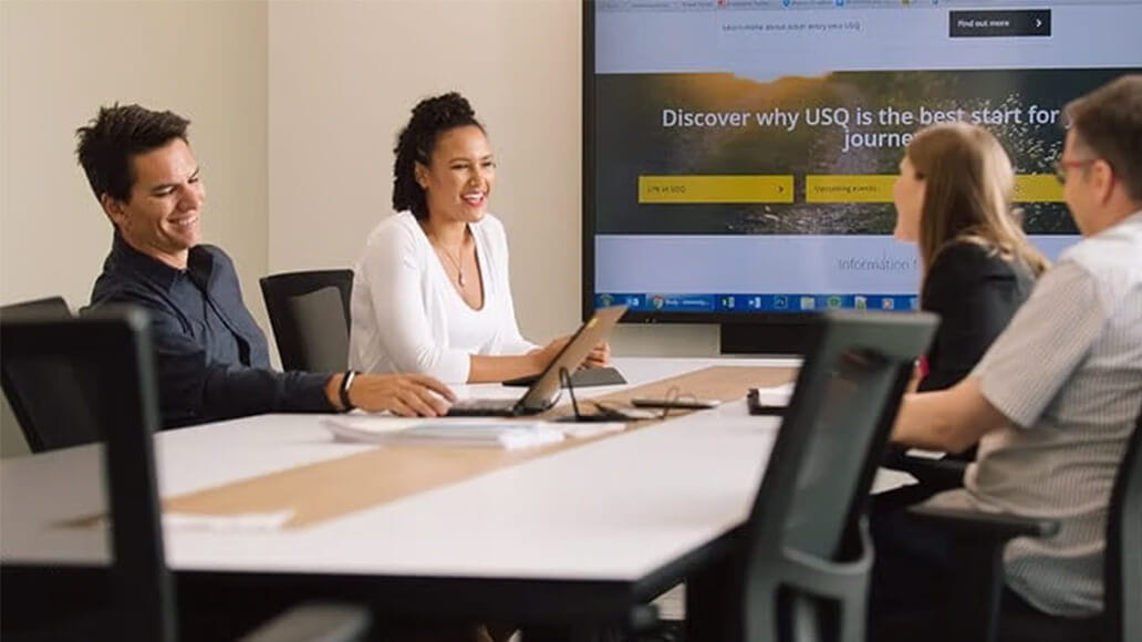 Four people are seated around a table in a meeting room, engaged in discussion with a screen displaying a website in the background.