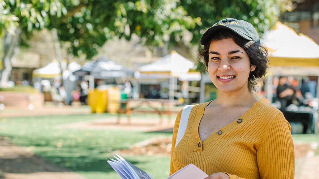 A person in a yellow sweater and black cap is holding papers and smiling in an outdoor setting with tents and trees in the background.