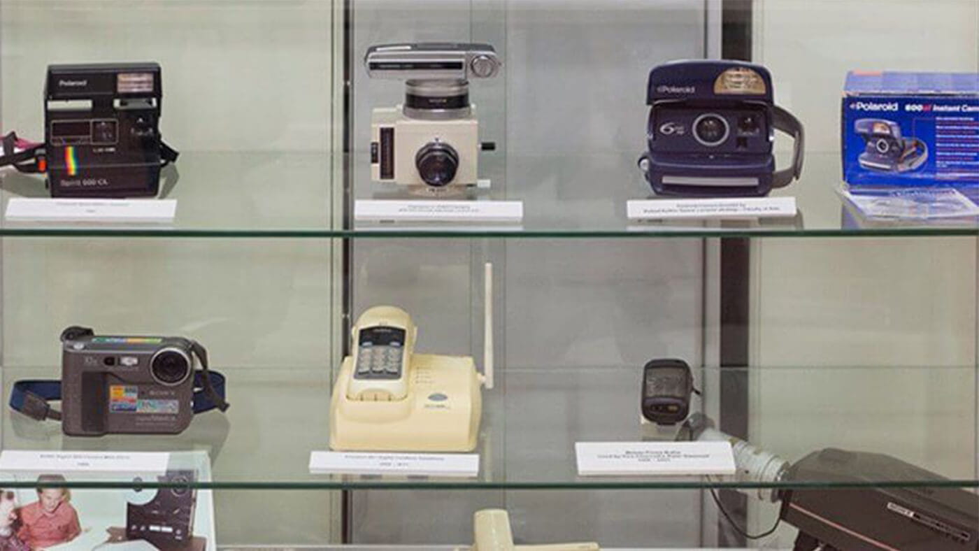 Glass display case showcasing various vintage Polaroid cameras, a cordless phone, and other electronic gadgets. Items are arranged on two shelves with labels indicating their respective models.