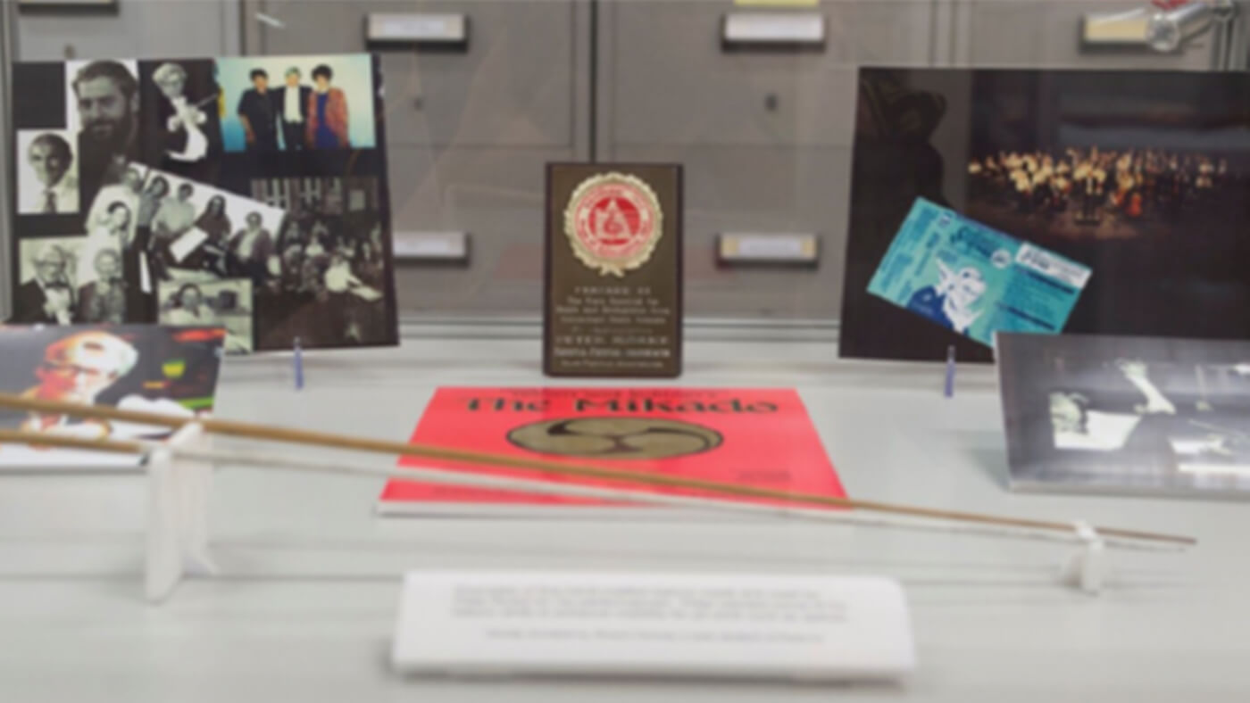 A display case featuring various photos, a plaque, a program for "The Mikado," and a conductor's baton.