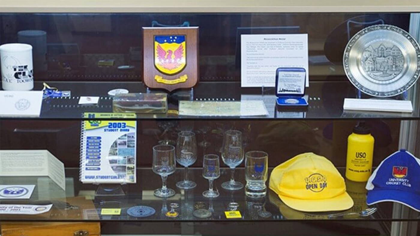 A display case exhibiting various memorabilia including a shield plaque, mugs, glassware, a plate, a calendar, a yellow cap, a medal, and informational cards.