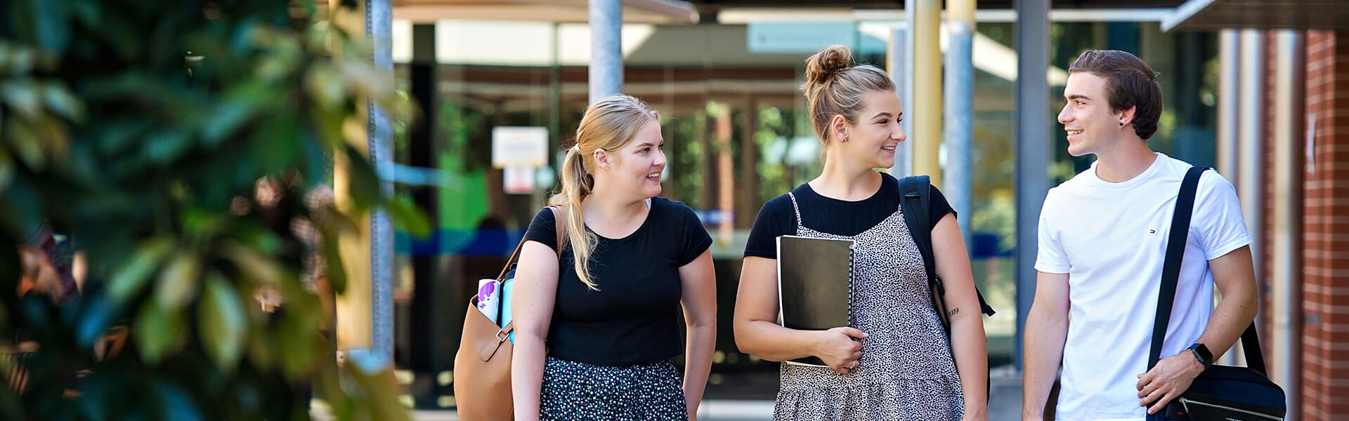 Three people walking and talking near a building entrance, each carrying bags and smiling.