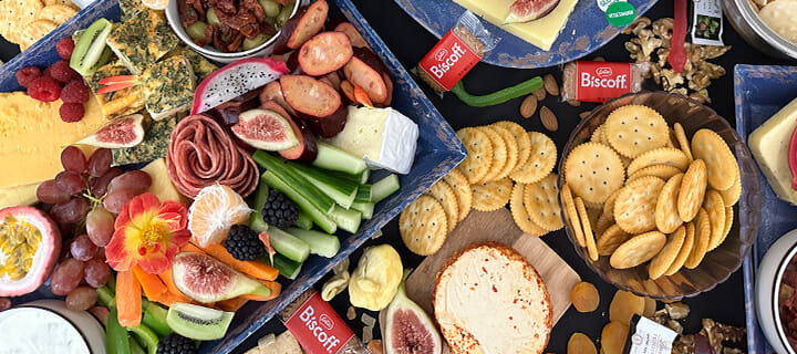 An assortment of cheese, crackers, figs, vegetables, and Biscoff cookies arranged on a platter.