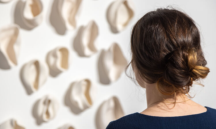 Person with brown hair in a bun observing an art installation of abstract, folded shapes on a wall.