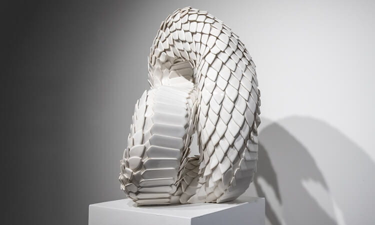 A textured, white abstract sculpture with curved, interlocking shapes is displayed on a pedestal against a gray background.