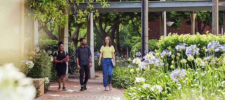 Three people walking on a brick path through a garden, surrounded by greenery and flowers.