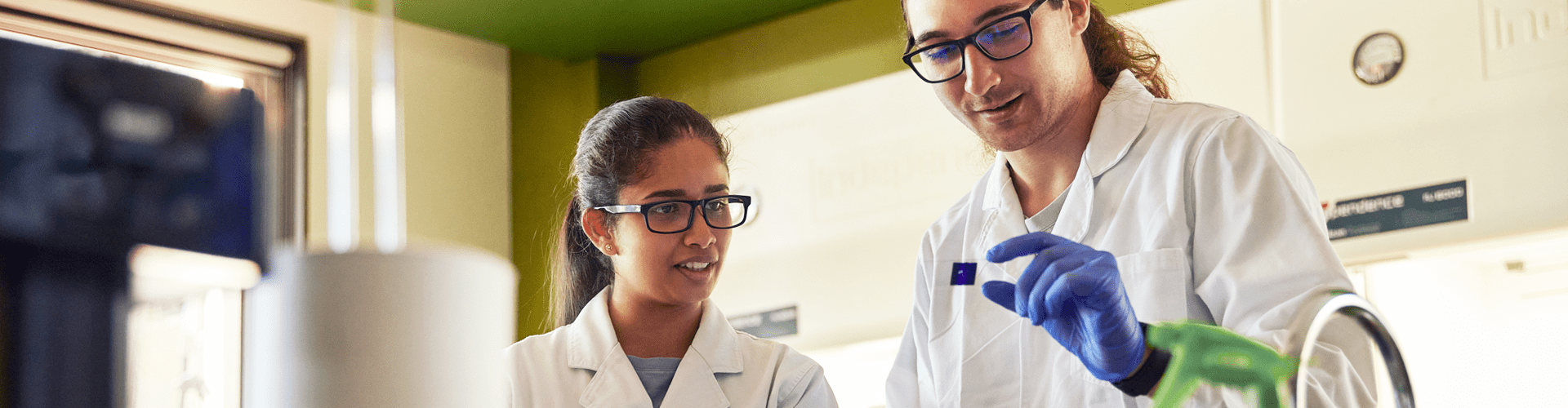 Two scientists wearing lab coats and gloves examine a slide in a laboratory setting.