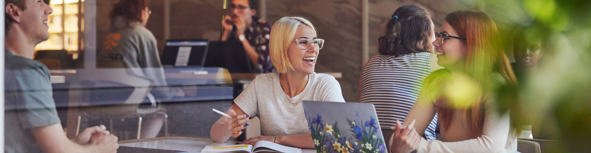 A group of people are sitting at a table, engaged in conversation and working on laptops in a casual setting.