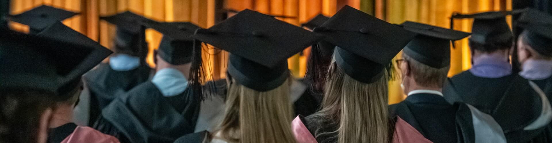 Graduates wearing caps and gowns sit facing a stage with gold curtains.