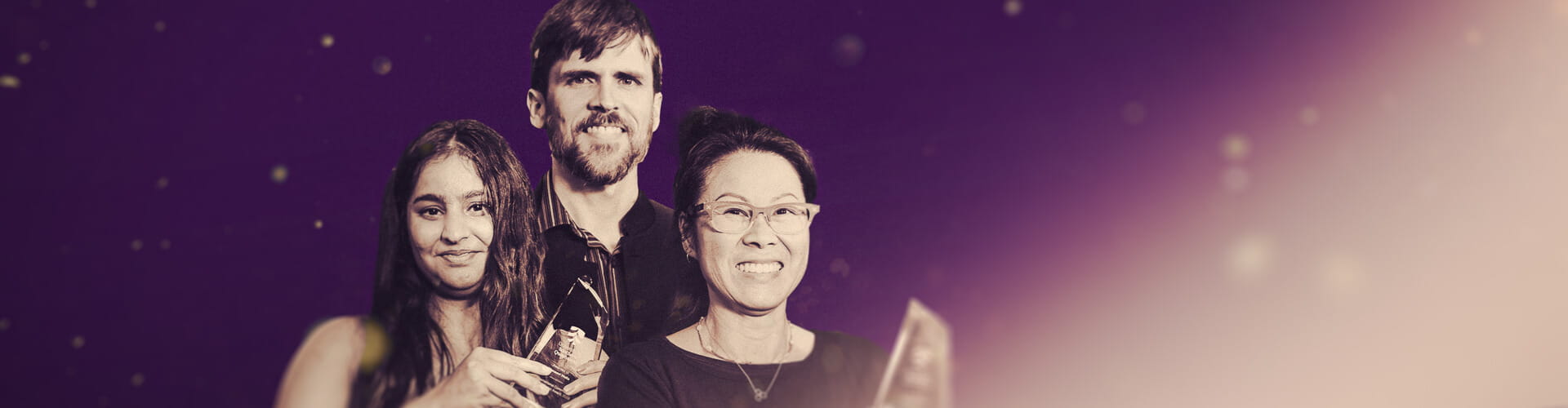 Three individuals smile while holding awards against a purple gradient backdrop with small glowing particles.