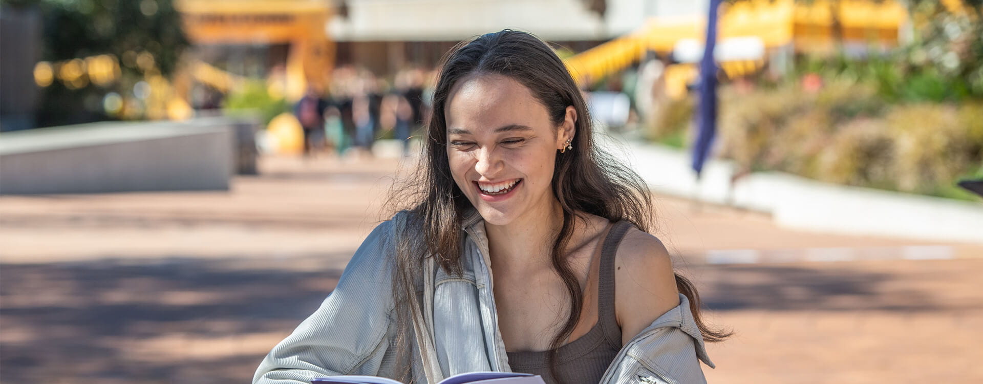 A person with long hair smiles while reading a book outdoors in a sunny area.