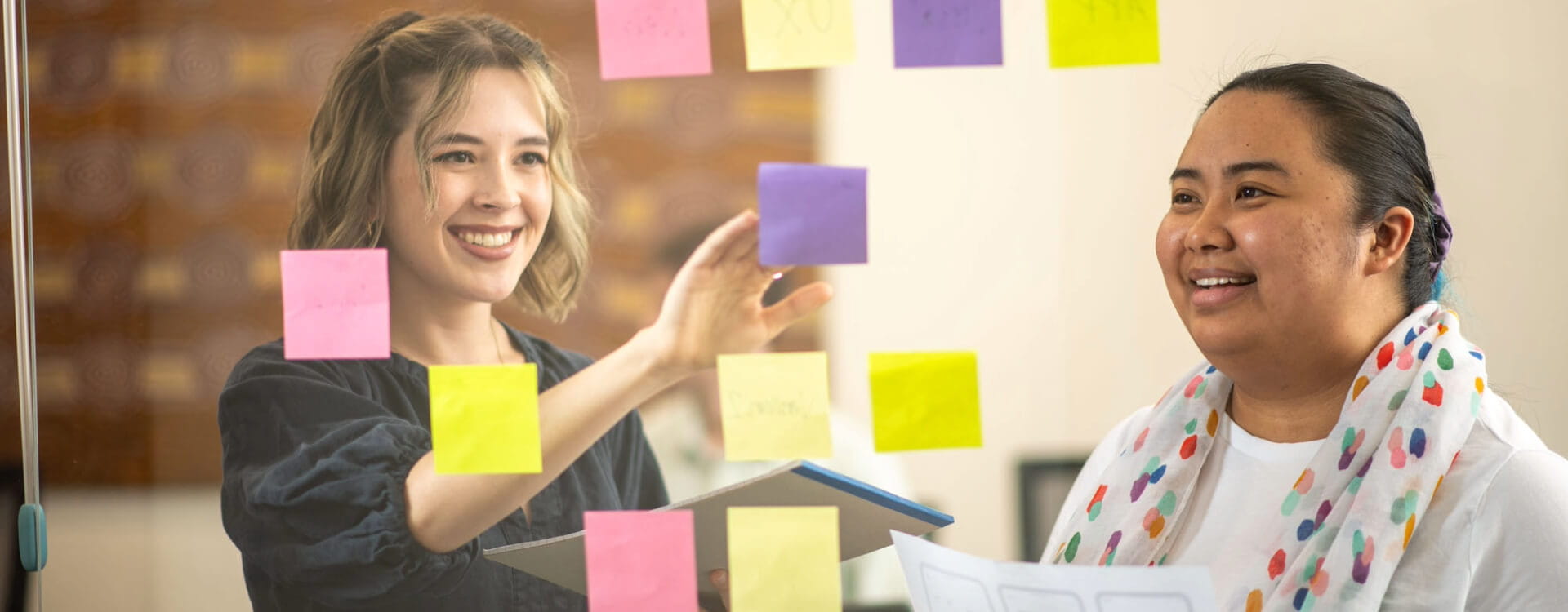 Two people in an office setting interact with colorful sticky notes on a transparent board, one smiling and pointing, the other holding documents.