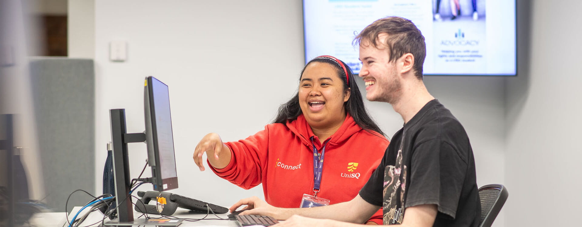 Two people at a desk using a computer, both smiling. The person on the left is pointing at the monitor and wearing a red shirt.