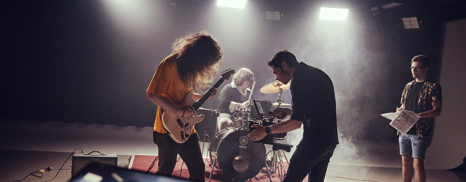 A band performs with a guitarist, drummer, and a cameraman filming. Another person stands nearby with a paper. Bright lights illuminate the scene.