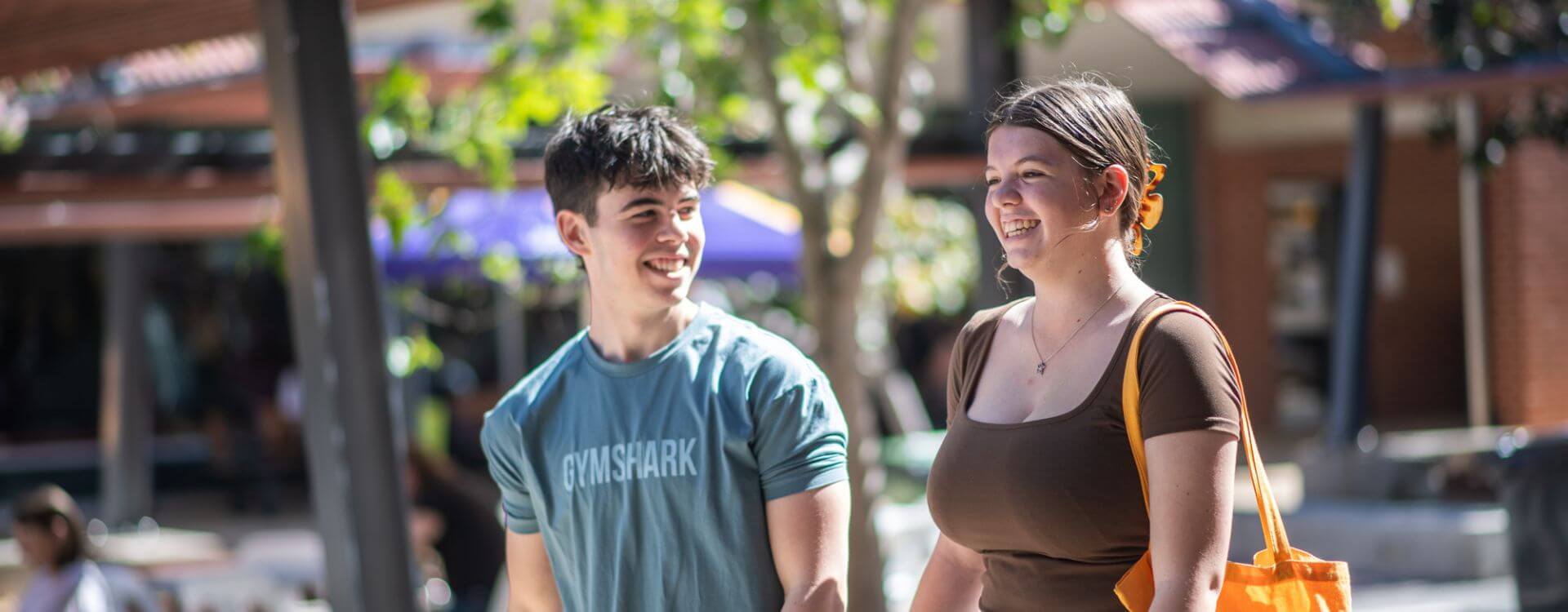 A young man and woman walk outdoors, smiling and talking. The man wears a light blue t-shirt, and the woman carries an orange bag. Trees and buildings are in the background.