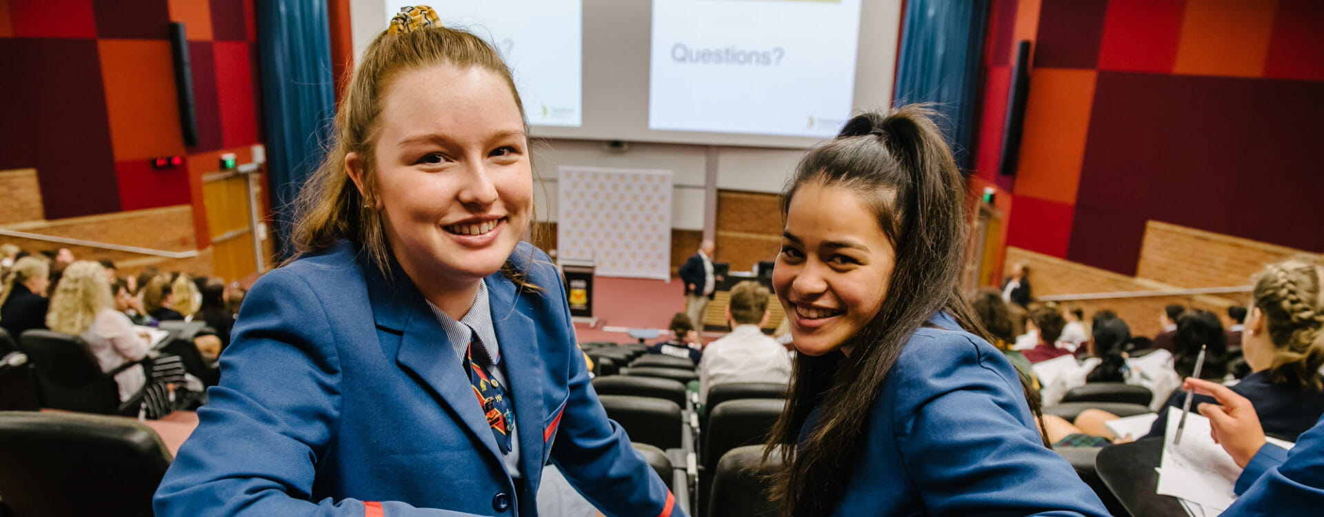 Two students in blue uniforms seated in a lecture hall, smiling at the camera. A presentation screen reads "Questions?" in the background. Audience members fill the room.