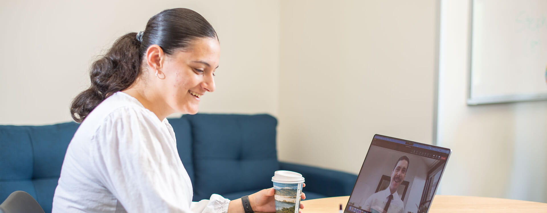 A woman holds a cup of coffee while video chatting on a laptop in an office setting.