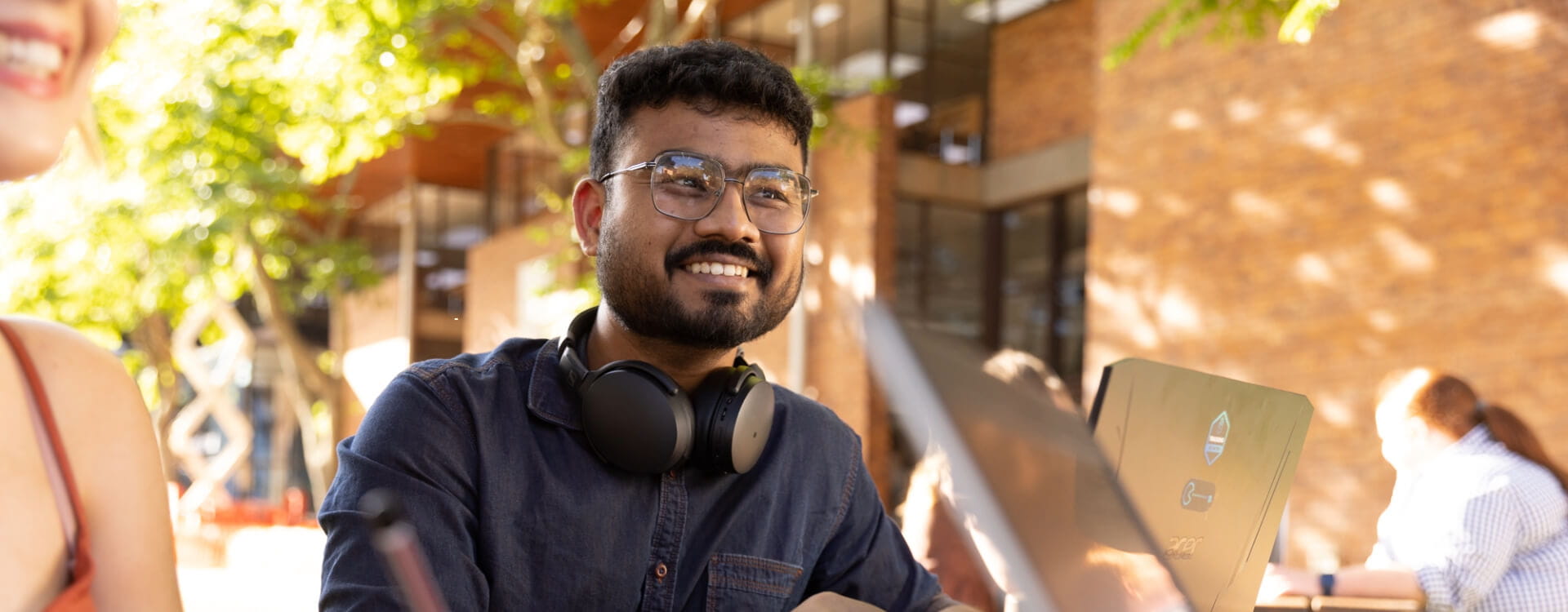 A person with glasses and headphones sits outdoors, smiling while looking at a laptop.