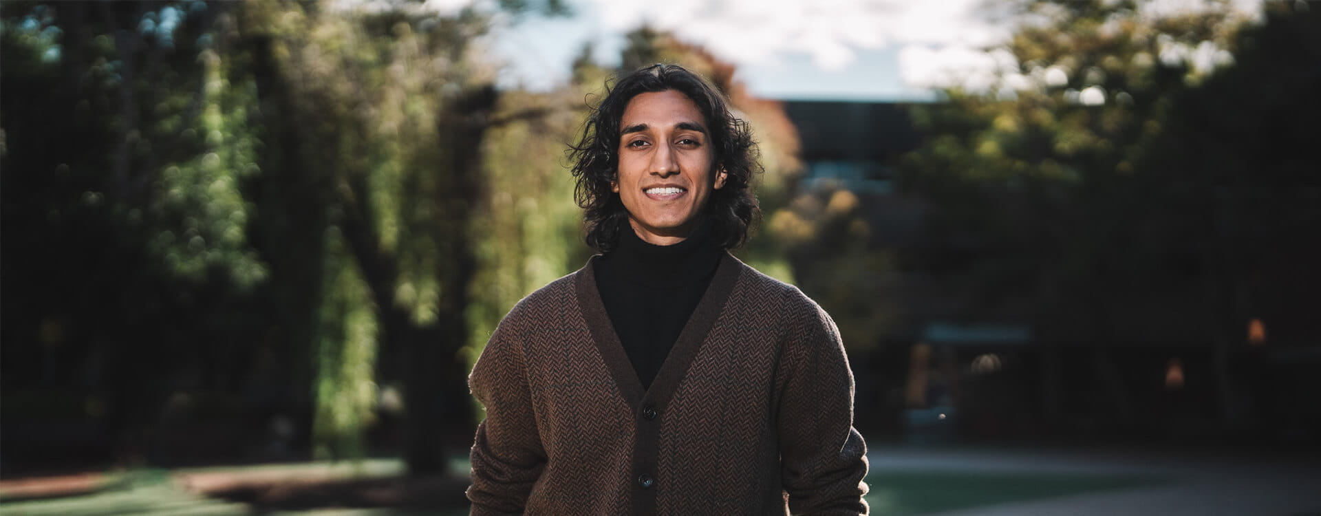 A person with long hair smiles outdoors, wearing a brown cardigan over a black shirt. Trees and sunlight are in the background.