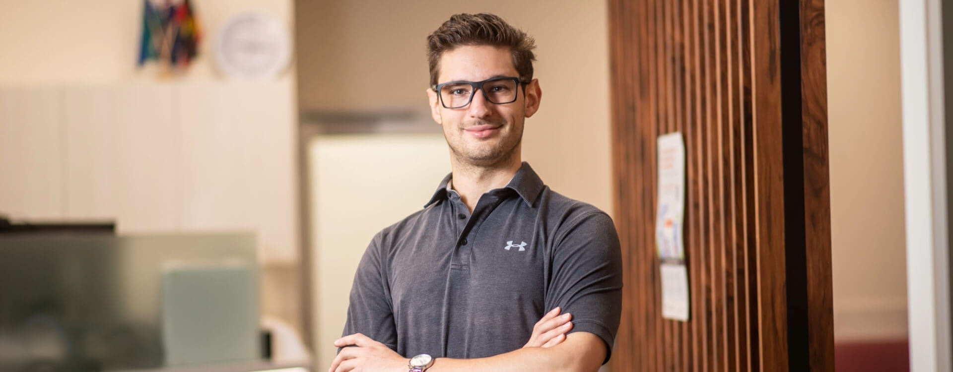 A man wearing glasses and a dark polo shirt stands with arms crossed in an indoor setting.