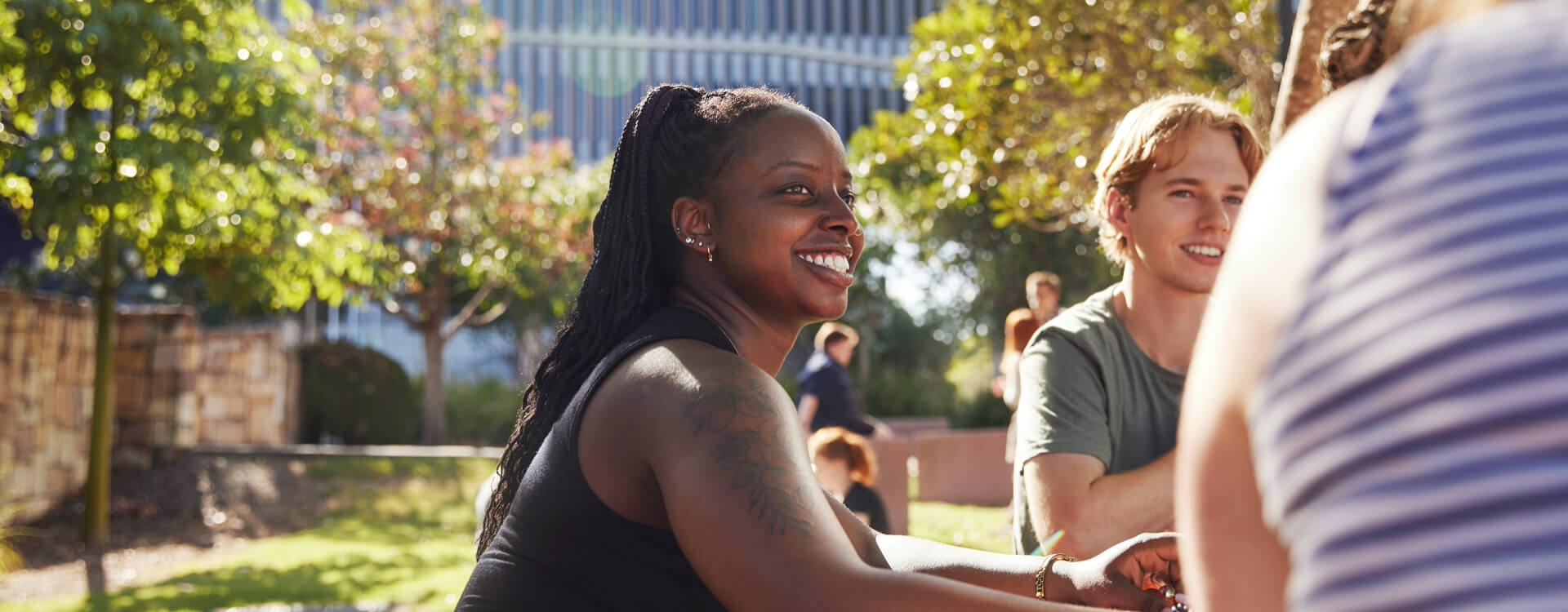 People sitting outdoors at a table, talking and smiling, with trees and buildings in the background.