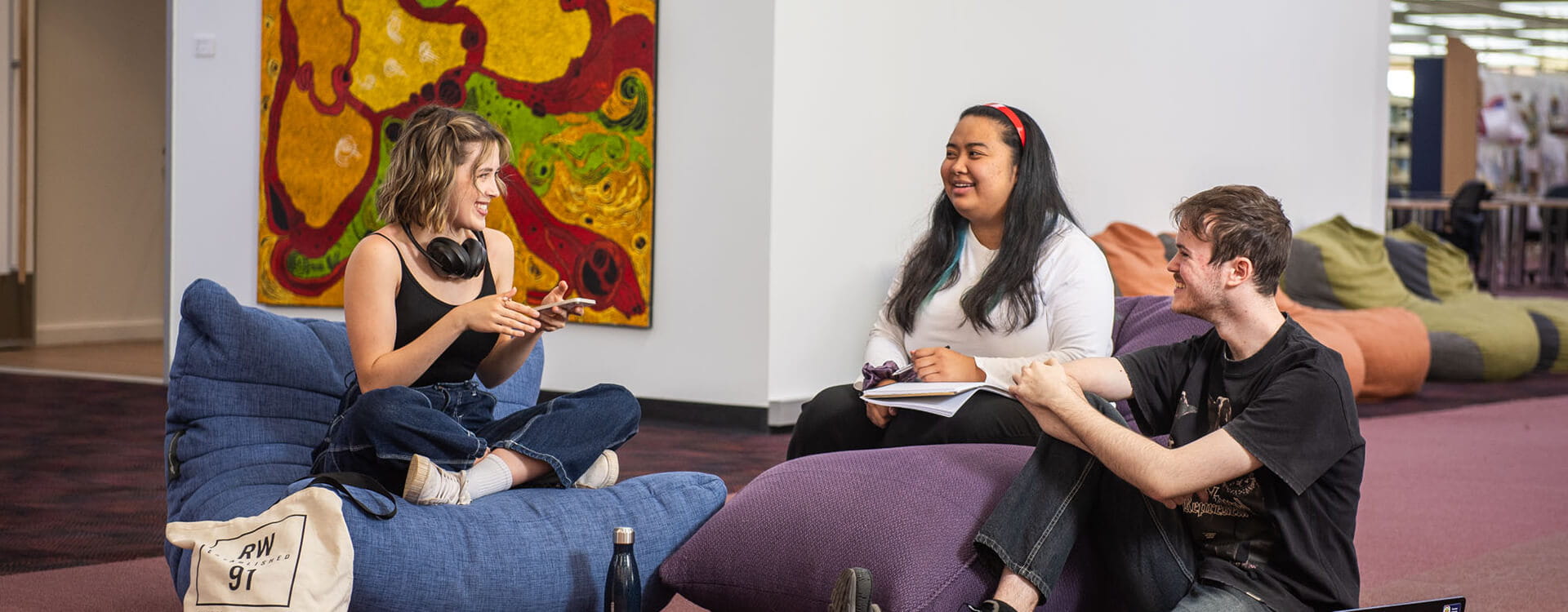 Three people sit on bean bags in a brightly lit room, engaged in conversation. A colorful abstract painting is on the wall behind them.