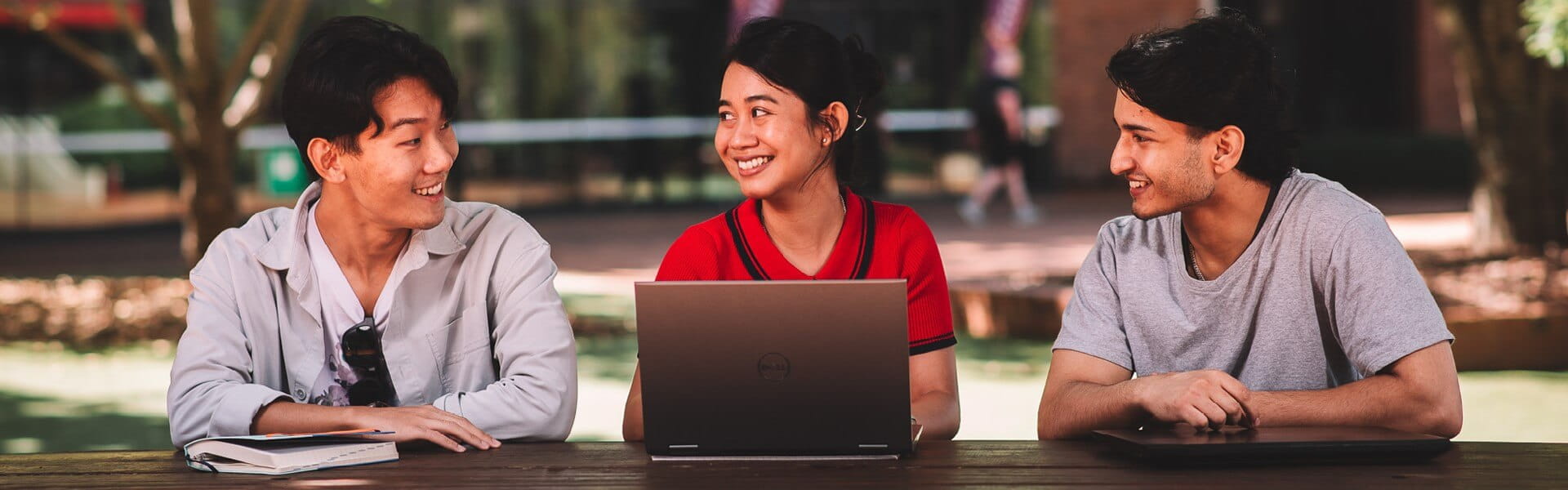 Three people sitting outdoors at a wooden table, smiling and looking at each other. Two laptops are present on the table.