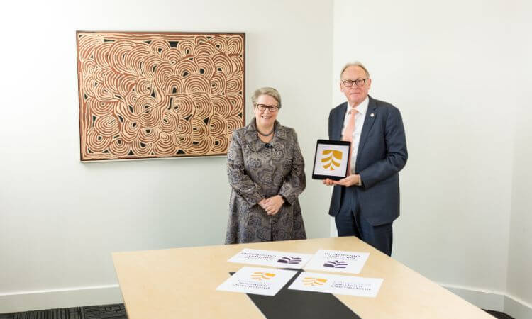 Two individuals standing in a room with an abstract painting in the background, exchanging an award or commemorative plaque.