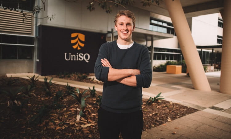 A person stands confidently with arms crossed in front of a building with a sign that reads UniSQ. 
