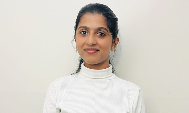 A person with long dark hair, wearing a white turtleneck top, stands against a plain white background.