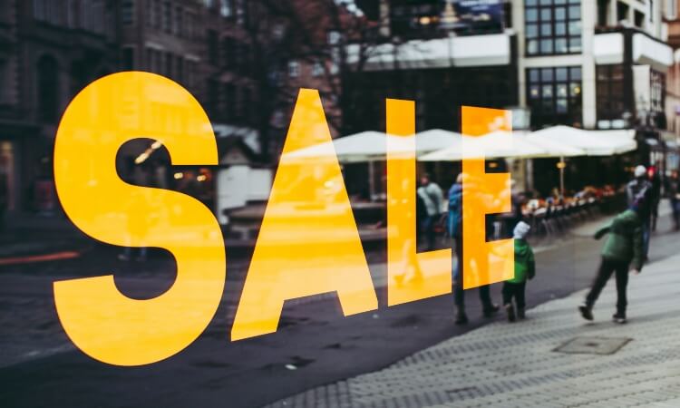 A yellow "SALE" sign is displayed on a storefront window with blurred people walking on the street outside.