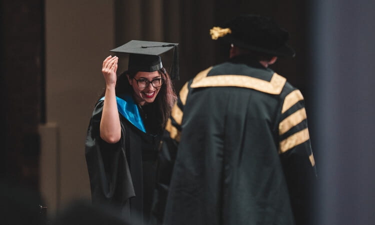 A graduate in a cap and gown smiles and adjusts her cap while walking past an official in ceremonial attire during a graduation ceremony.