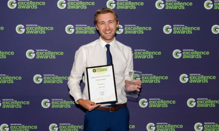 A man in a white shirt and blue tie holds a certificate and a glass award in front of a backdrop reading "Geospatial Excellence Awards".
