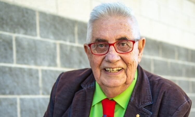 A man wearing red glasses, a green shirt, a colorful tie, and a brown jacket smiles in front of a gray brick wall.