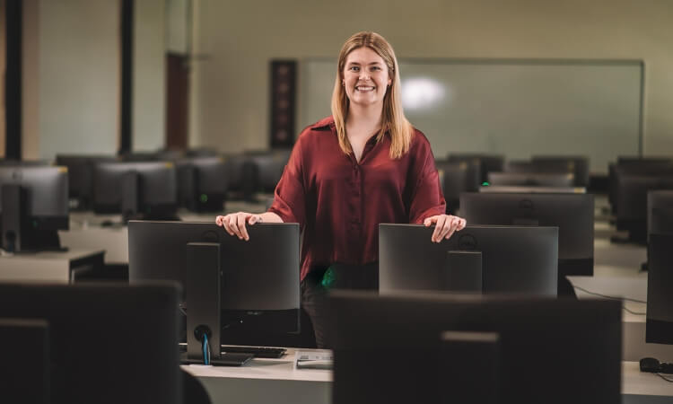 A woman in a red blouse stands in a computer lab, smiling, with her hands on the back of a chair in front of a monitor, surrounded by multiple workstations.