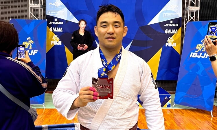A man holds a medal and poses for a photo with a backdrop of a martial arts competition. 