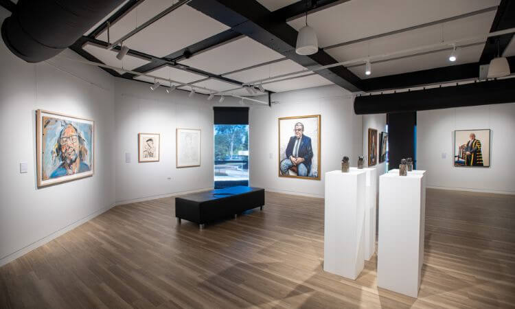 A modern art gallery interior with wooden floors showcasing portraits on white walls and sculptures on white pedestals. A black bench is centrally placed in the room.