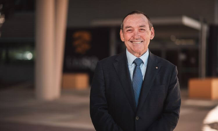 An older man with a receding hairline, dressed in a dark suit and blue tie, smiles while standing outside a modern building.