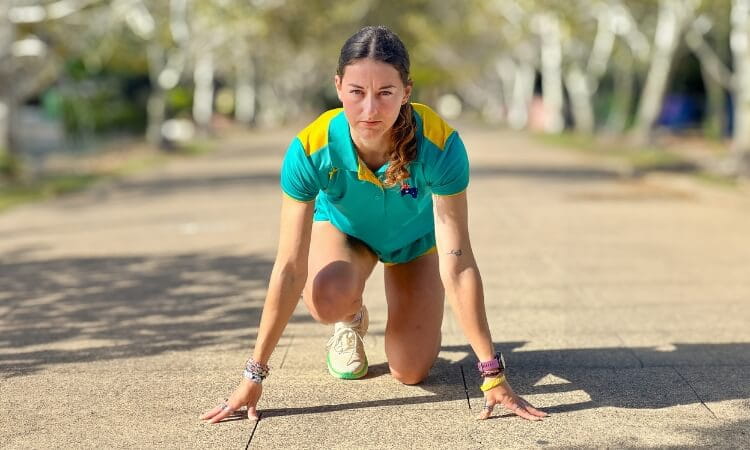 A person in athletic attire is crouched on a paved pathway in a starting position, preparing to run. 