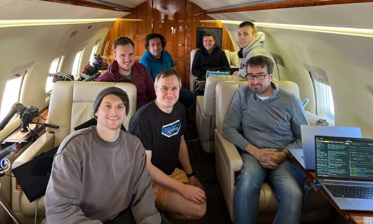 Seven people are seated inside a plane with laptops and equipment visible. 