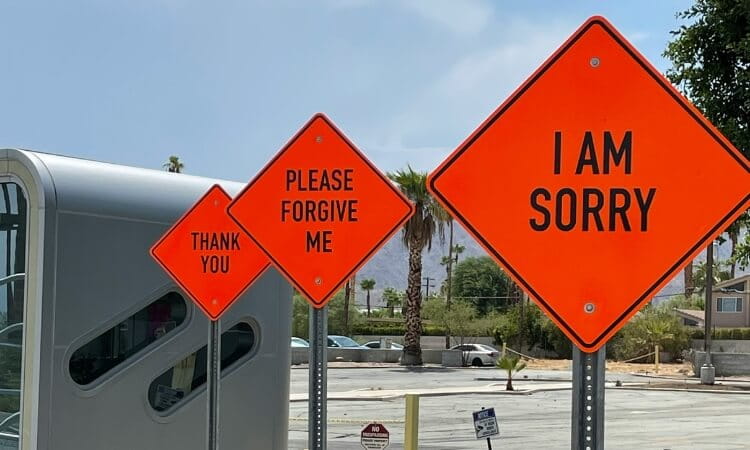 Three orange diamond-shaped signs with black text read "THANK YOU," "PLEASE FORGIVE ME," and "I AM SORRY" in a row on metal poles in an outdoor setting with trees and buildings in the background.